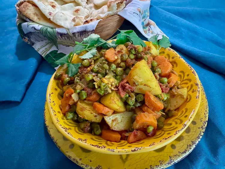 A vibrant bowl of vegetable curry featuring potatoes, carrots, and peas, garnished with fresh cilantro, served with naan bread.